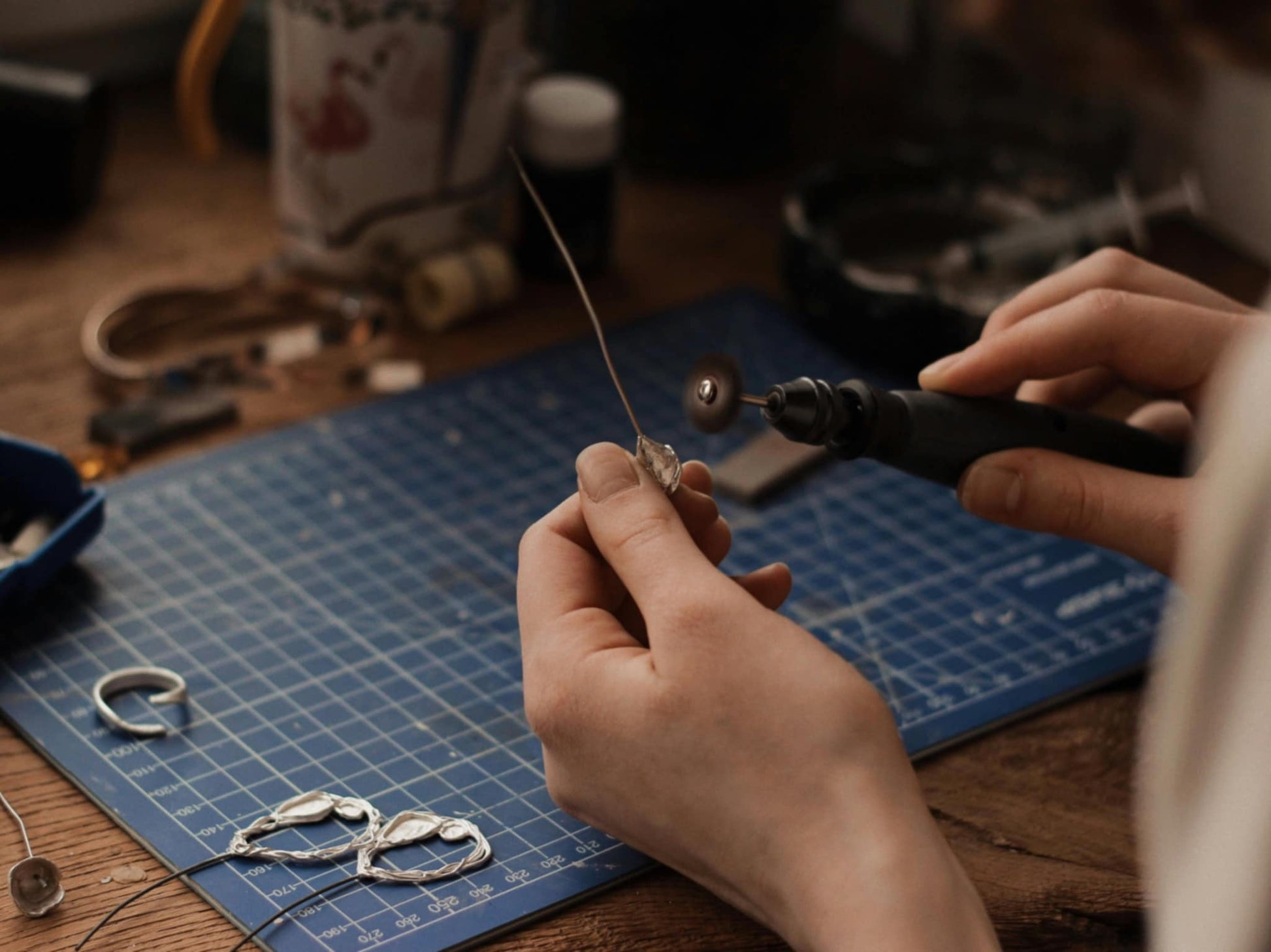 A close-up of a jeweler’s hands using a rotary tool to polish a handcrafted piece. The workspace features a cutting mat, tools, and partially finished jewelry, showcasing the delicate process of jewelry making.