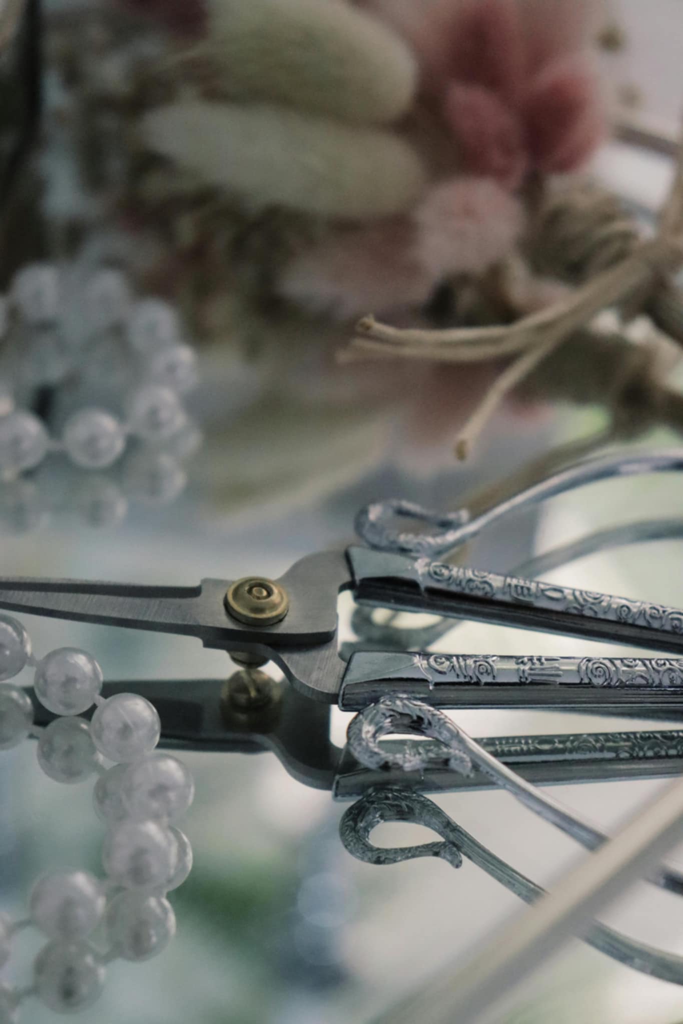 
 A close-up of jewelry-making tools with intricate engravings, delicate beads, and soft dried flowers in the background. The scene reflects the artistry and precision of handcrafted jewelry.